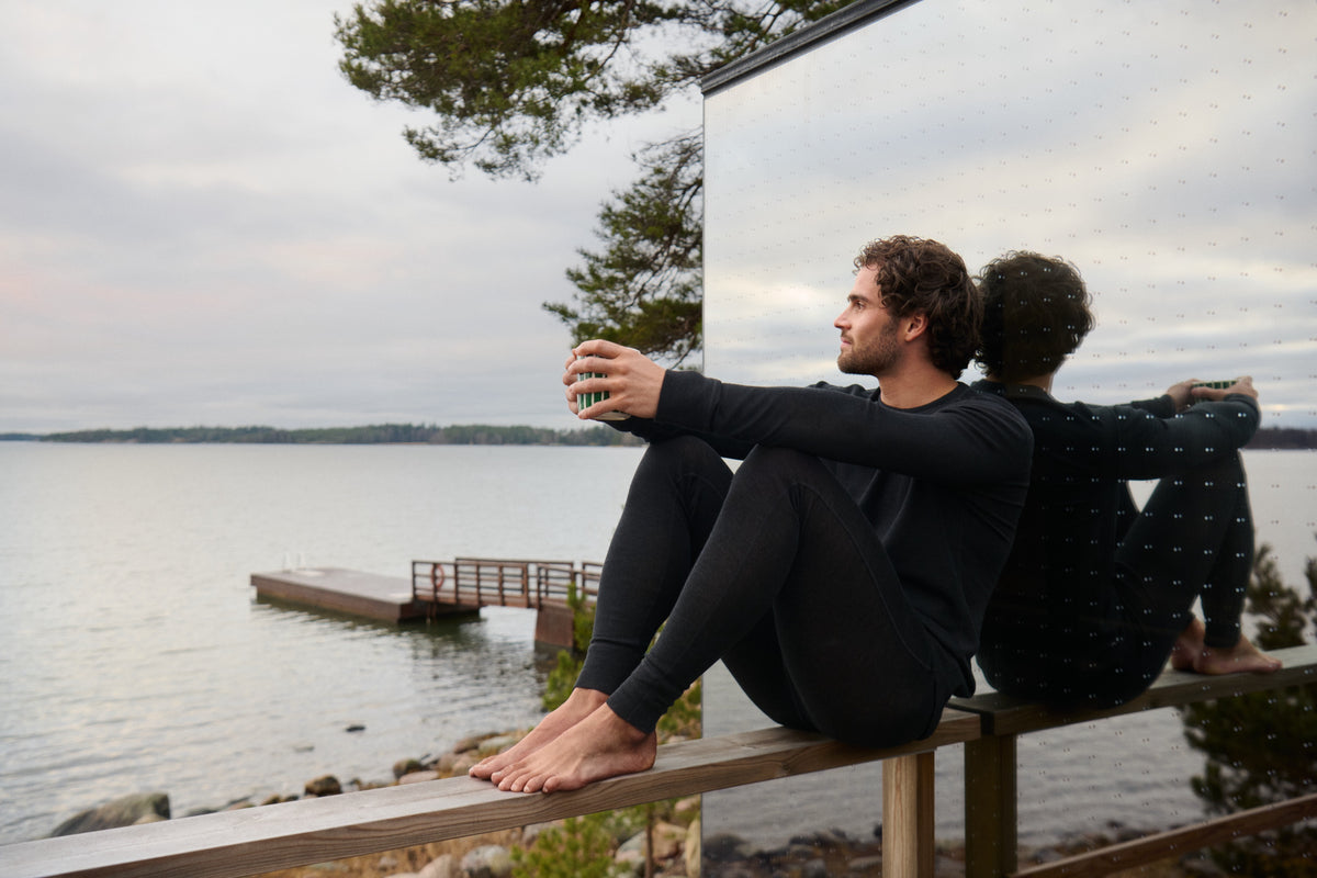 Mies istuu järven rannalla puukaiteella mustassa kerrastossa kuppi kädessään.